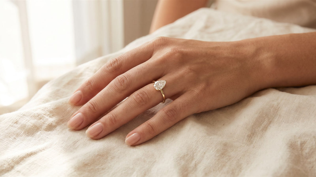 Woman's hand resting on cream linen wearing a pear shaped diamond ring in yellow gold
