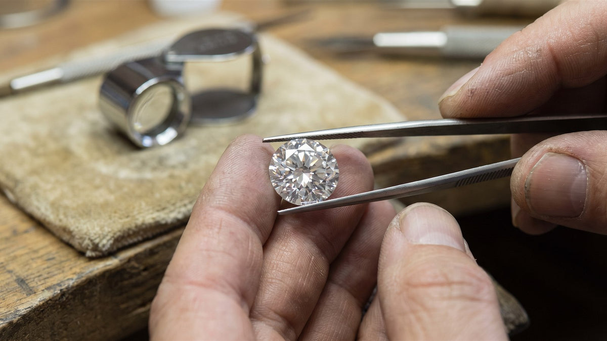 A jeweller holds a round brilliant diamond in tweezers above a workbench with a loupe